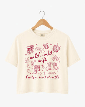 Bachelorette weekend crop top with Wild Wild Wife theme, playful line-art doodles, and script lettering. Personalized with bride name, EST year, and destination city for the whole crew.