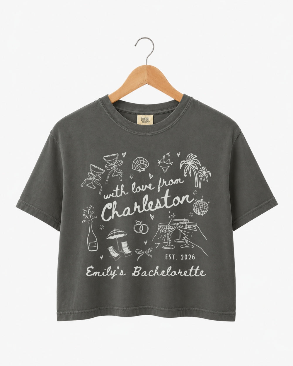 Cropped bachelorette top with hand-drawn doodle script and playful icons, Charleston weekend theme. Personalized with bride name, destination city, and EST year for the whole crew.
