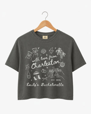Cropped bachelorette top with hand-drawn doodle script and playful icons, Charleston weekend theme. Personalized with bride name, destination city, and EST year for the whole crew.